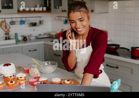 Sorridente giovane panettiera che usa il computer portatile e parla al telefono mentre prepara torte in cucina Foto Stock