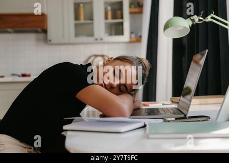 Giovane donna d'affari stanca che si appoggia al laptop e dorme mentre si siede alla scrivania a casa Foto Stock