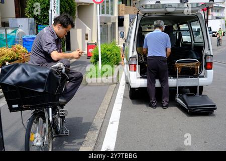 Un messaggero di biciclette seduto sulla sua bicicletta e guardando il suo telefono quando si prende una pausa con il personale addetto alla consegna e il suo furgone sullo sfondo. Harajuku, Shibuya Ward, Tokyo Foto Stock