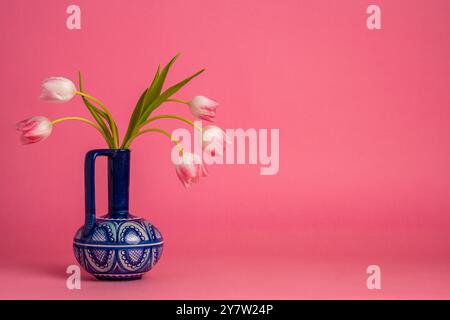 Tulipani rosa tenui in vaso blu su sfondo rosa. I fiori delicati si armonizzano con lo sfondo luminoso, mentre il vaso scuro aggiunge contrasto alla composizione. Ancora Foto Stock