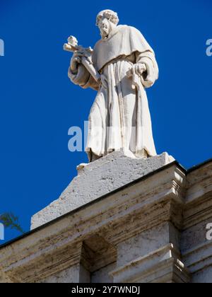 Mantova, Italia 30 settembre 2024 un'antica statua intemprata di un santo che regge un crocifisso in cima ad una chiesa contro un cielo blu Foto Stock