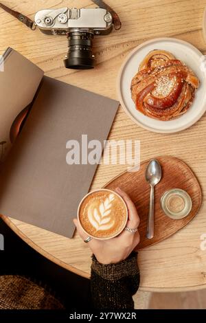 Una donna tiene il caffè con le mani su un tavolo in una caffetteria, circondata da deliziosi pasticcini, riviste e una macchina fotografica. Vista dall'alto. Foto Stock