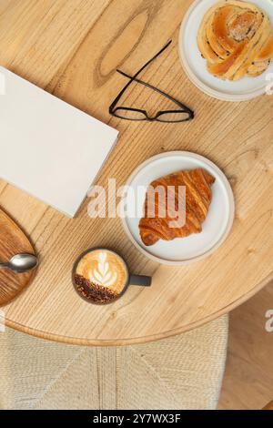 Un tavolo in una caffetteria, con una tazza di cappuccino, deliziosi pasticcini e riviste. Vista dall'alto. Foto Stock