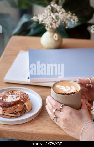 La donna tiene il caffè con le mani su un tavolo in una caffetteria, circondata da deliziosi pasticcini e riviste. Foto Stock