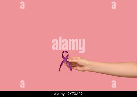 Mano femminile con nastro viola su sfondo rosa. Concetto di consapevolezza del cancro al pancreas Foto Stock