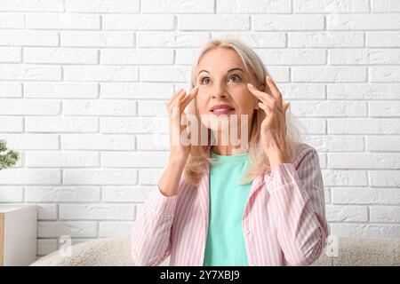 Donna matura che tocca le rughe vicino agli occhi contro il muro di mattoni chiari Foto Stock