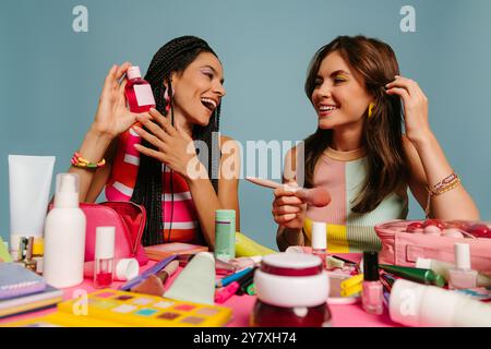Due influencer donne felici che esaminano vari prodotti di bellezza mentre sono sedute alla scrivania su sfondo blu Foto Stock