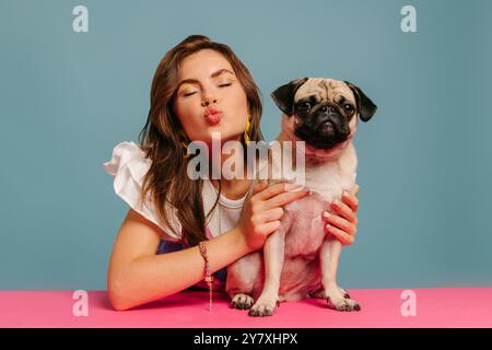 Bella donna che soffia come bacio e abbraccia un carino cane mentre siede alla scrivania rosa su sfondo blu Foto Stock