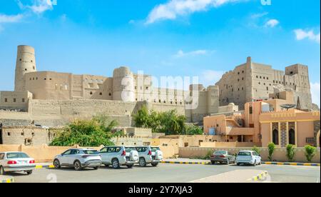 Fortezza della cittadella di Bahla, mura di pietra e torri rotonde con vecchio edificio e parcheggio pieno di auto in primo piano, Bahla, Oman Foto Stock