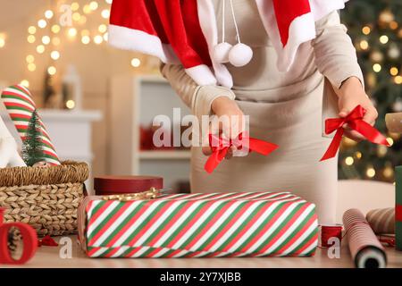 Bella giovane donna a Santa cape che avvolge la confezione regalo di Natale con arco sul tavolo a casa, primo piano Foto Stock