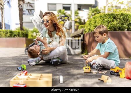 Mamma e figli giocosi che si divertono seduti a terra e giocano con i giocattoli in un parco Foto Stock