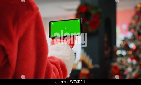 L'assistente del personale che indossa la tuta di Babbo Natale nel centro commerciale adornato con decorazioni natalizie che tengono il telefono con chiave cromatica, prendendosi una pausa dal lavoro mentre guardi i video sullo schermo verde, primo piano Foto Stock