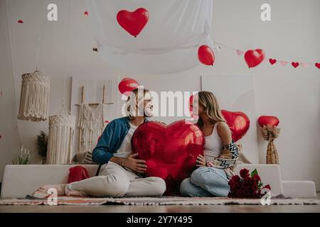 Coppia felice che tiene in mano un palloncino a forma di cuore rosso mentre festeggia San Valentino in un'accogliente casa Foto Stock