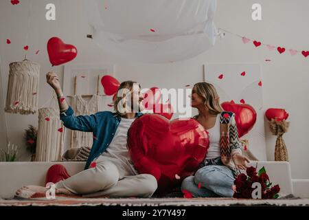 Felice coppia amorevole che tiene in mano un palloncino a forma di cuore rosso e lancia coriandoli mentre festeggia San Valentino a casa Foto Stock