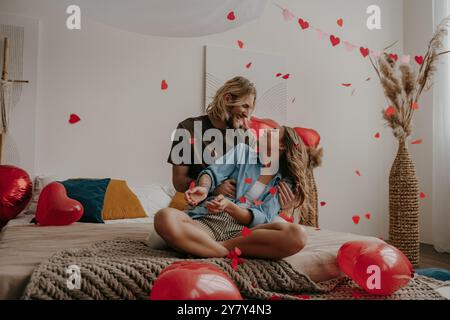Felice coppia amorevole seduto sul letto e gettando coriandoli a forma di cuore mentre festeggiano San Valentino a casa Foto Stock