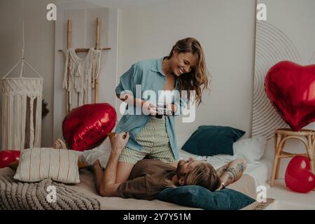 Bella giovane donna sorridente che fotografa il suo fidanzato con una fotocamera retrò mentre entrambi si rilassano a letto a casa Foto Stock