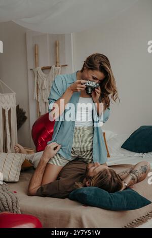 Bella giovane donna che fotografa il suo fidanzato con una fotocamera retrò mentre si rilassa a letto a casa Foto Stock