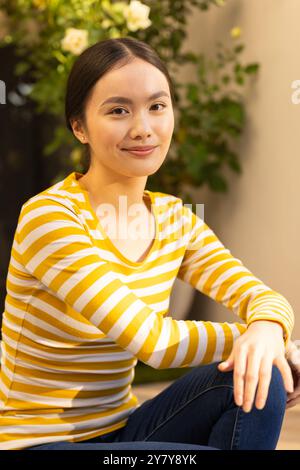 Sorridente ragazza asiatica in camicia a righe gialle seduta all'aperto, guardando la macchina fotografica Foto Stock