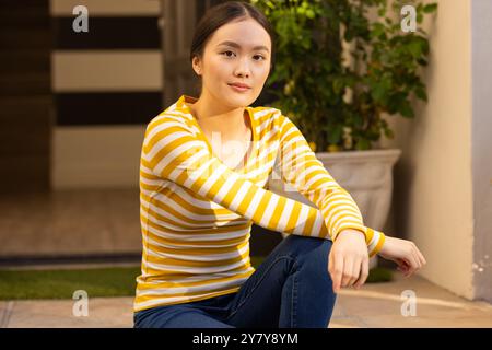 Sorridente ragazza asiatica in camicia a righe seduta all'aperto, godendo di una giornata di sole Foto Stock