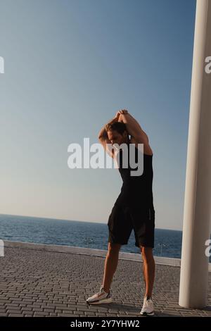 Giovane forte in abbigliamento sportivo che fa esercizi mattutini con il mare sullo sfondo Foto Stock