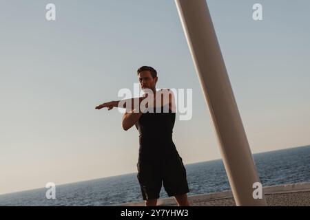 Giovane atletico in abbigliamento sportivo che fa esercizi di stretching con il mare sullo sfondo Foto Stock