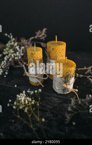 Tre candele di cera d'api artigianali con una consistenza a nido d'ape, adornate da fiori secchi e spago, si trovano su una superficie scura. Foto Stock