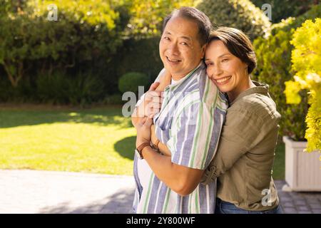 Una coppia, una donna sorridente di mezza età che abbraccia un uomo asiatico all'aperto in un giardino soleggiato, godendosi il tempo libero in famiglia Foto Stock