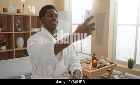 Bell'uomo che scatta selfie al chiuso nel centro benessere con accappatoio circondato da arredi benessere e articoli di bellezza Foto Stock