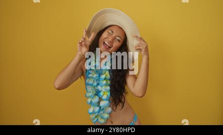 Bella giovane donna che posa su uno sfondo giallo con un cappello e lei hawaiana, con un giocoso segno di pace e un sorriso, che irradia vibrazioni estive a. Foto Stock