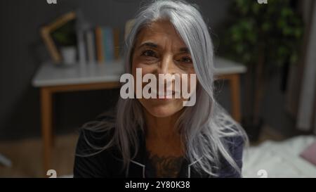 Donna sorridente di mezza età al coperto in camera da letto dai capelli grigi seduto sul letto matura donna caucasica casa bella Foto Stock