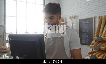 Giovane che lavora in una panetteria guardando uno schermo di un computer con pane fresco sullo sfondo, indossando un grembiule e stando in piedi in un luminoso negozio al coperto Foto Stock