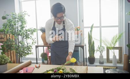 Giovane asiatico che usa lo smartphone mentre organizza fiori in un luminoso negozio di fiori al coperto. Foto Stock