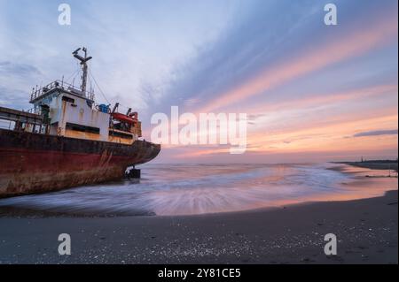 Una nave intemprata si staglia contro un cielo infuocato al tramonto sulle sabbie dorate di Xigushui Beach. Il contrasto tra il cielo vibrante e l'oscurità Foto Stock