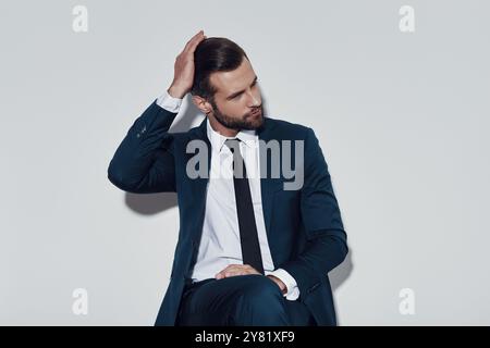 Momento di pensare. Bel giovane che regola la sua acconciatura seduto su uno sfondo grigio Foto Stock