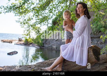 Donna in abito bianco e suo figlio seduto su un albero caduto in riva al lago, circondato dal verde in una giornata di sole. Foto Stock