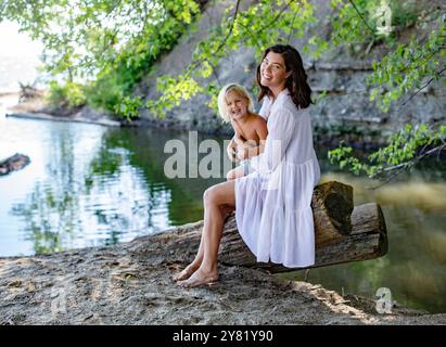 Donna in abito bianco e suo figlio seduto su un albero caduto in riva al lago, circondato dal verde in una giornata di sole. Foto Stock