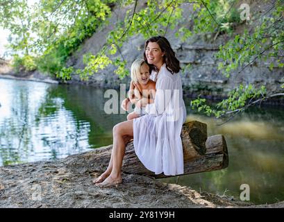 Donna in abito bianco e suo figlio seduto su un albero caduto in riva al lago, circondato dal verde in una giornata di sole. Foto Stock