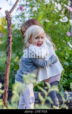 Una giovane ragazza gioiosa con i capelli biondi che viene abbracciata da un altro bambino in un vivace giardino. Foto Stock