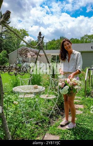 Donna sorridente in piedi in un lussureggiante giardino che organizza un bouquet di fiori accanto ai mobili da giardino vintage sotto un cielo limpido e soleggiato. Foto Stock