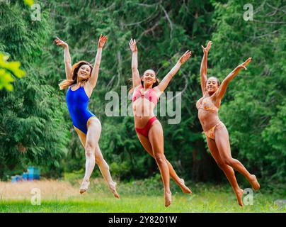 Tre donne gioiose in costume da bagno saltano in aria contro uno sfondo di verde. Foto Stock