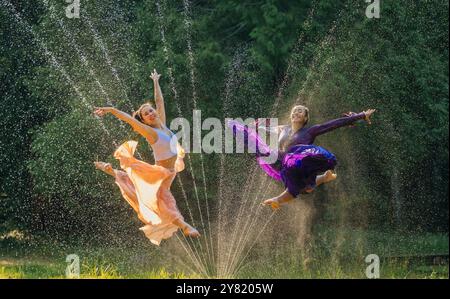 Due ballerini in costumi colorati saltano attraverso uno spruzzo d'acqua su un lussureggiante sfondo verde Foto Stock