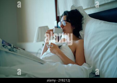 Una donna si rilassa a letto con una tazza di caffè e un quotidiano in un'accogliente camera da letto. Foto Stock