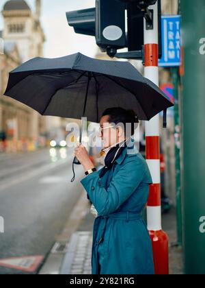 Una donna sorridente con un cappotto blu tiene un ombrello aperto mentre si trova in piedi su un passaggio pedonale. Foto Stock