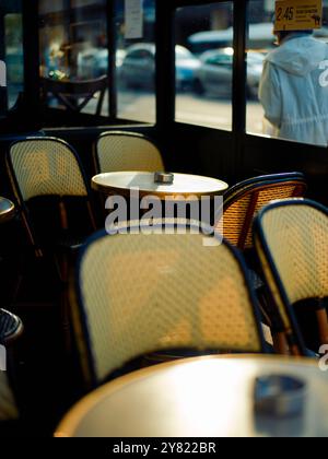 La luce del sole filtra attraverso una finestra che illumina l'interno di un caffè vuoto con tavoli rotondi e sedie in vimini, creando un'atmosfera calda e invitante. Foto Stock