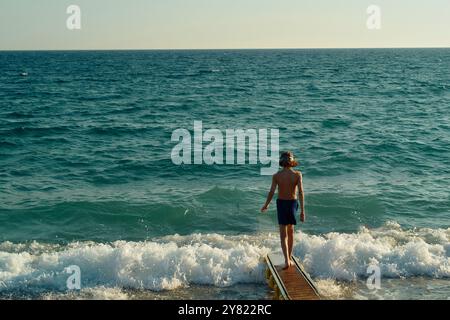 Un uomo a piedi nudi si trova alla fine di un molo di legno che guarda alle onde di un mare blu. Foto Stock