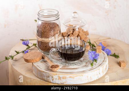 Composizione di una tazza di bevanda a base di cicoria, barattoli di cicoria solubile granulata e zucchero di canna, cucchiaio su un ceppo di legno. La cicoria è decaffeinato bere sana emozione Foto Stock