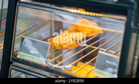 Vista ravvicinata dei pezzi di pollo fritti esposti in una vetrina di vetro. Il pollo è dorato e croccante, appoggiato su una griglia metallica. Le etichette dei prezzi sono Foto Stock