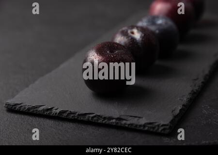 Succosa prugna fresca matura con gocce d'acqua sul tabellone in ardesia nero. Frutta di stagione su sfondo nero. Messa a fuoco selettiva, primo piano. Foto Stock