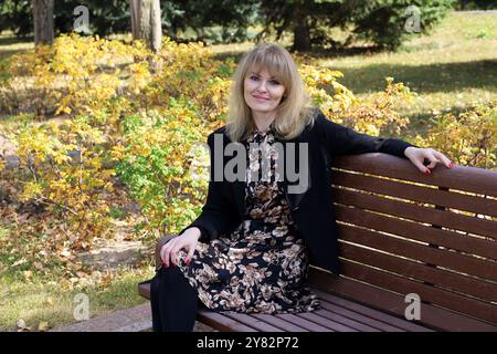 Ritratto all'aperto di una bella ragazza caucasica sorridente con capelli biondi seduta su una panchina nel parco autunnale Foto Stock