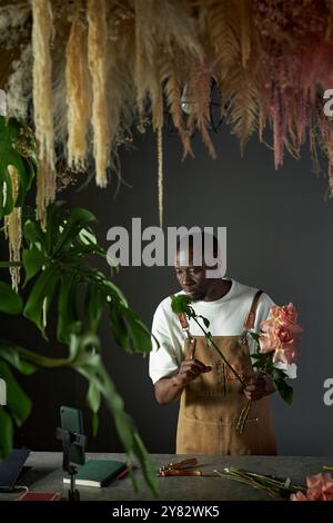 Ritratto verticale di un giovane fioraio maschio che crea un bouquet di fiori e registra video per i social media in un ambiente elegante e buio Foto Stock